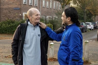 Twee mannen, een oudere en een jongere, staan buiten en lachen met elkaar, met bomen en auto's op de achtergrond.